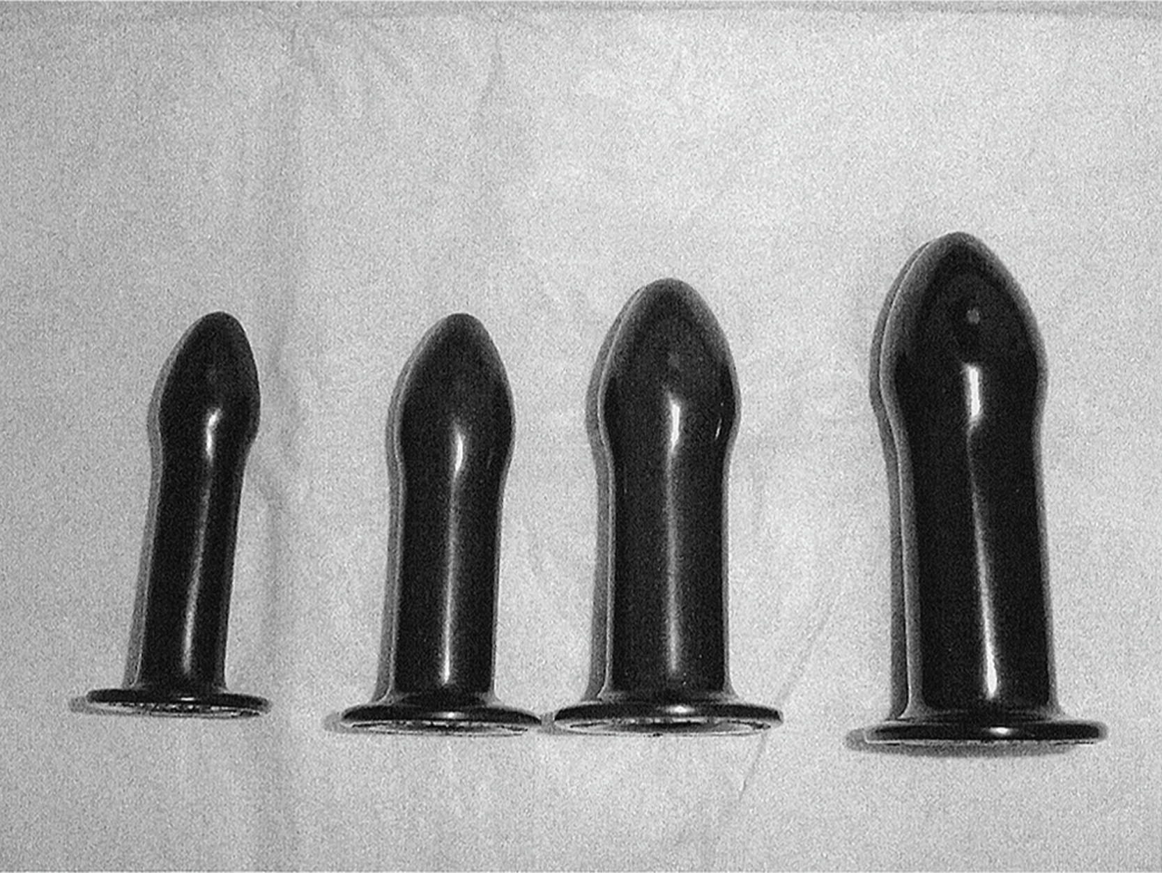 A clinical image of four cylindrical vaginal dilators, with rounded, slightly tapered tip and flared base. The size of dilators increases progressively from left to right in both length and diameter